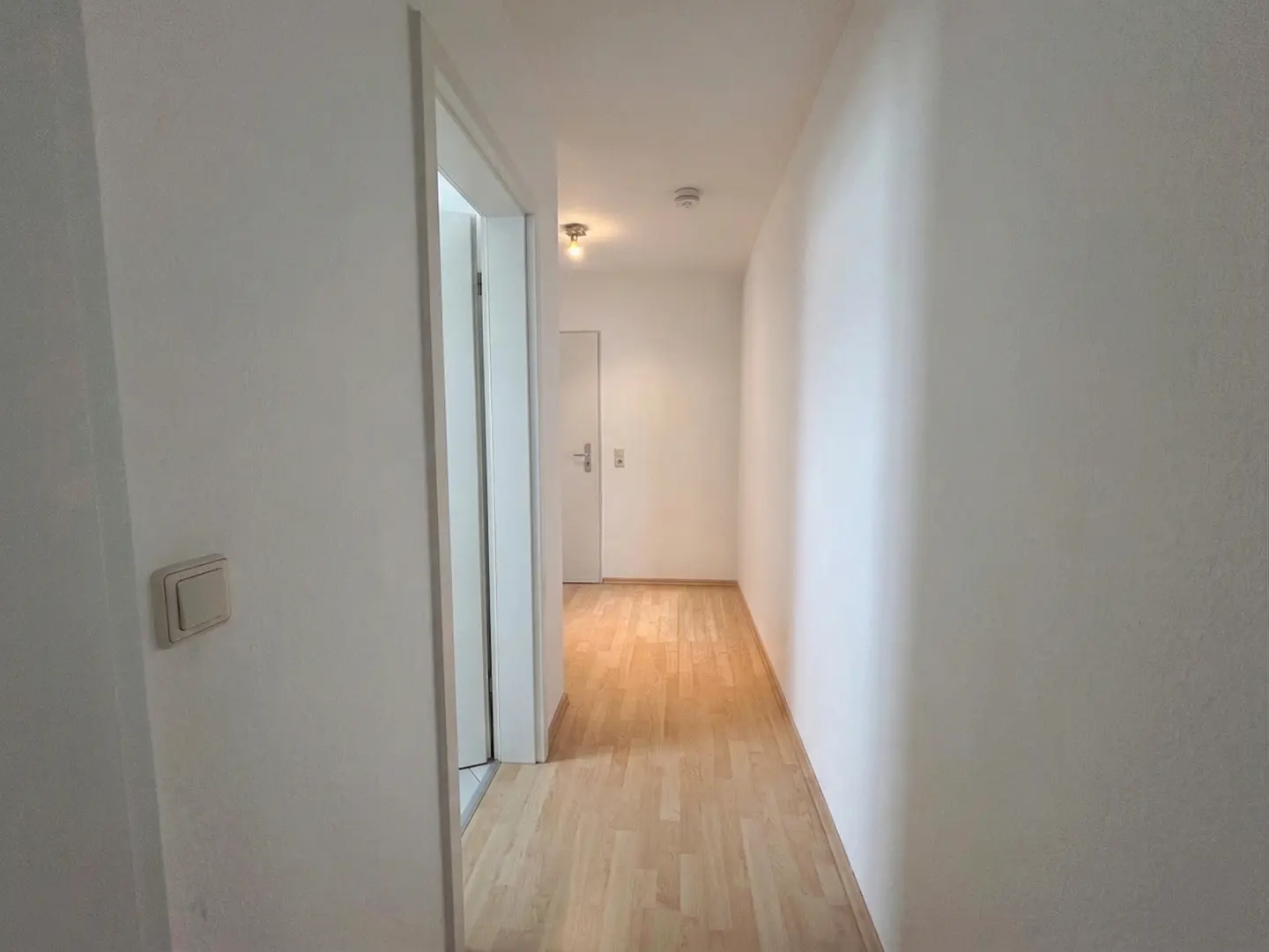 A bright hallway with white walls and light wood flooring leads to a closed white door at the end.