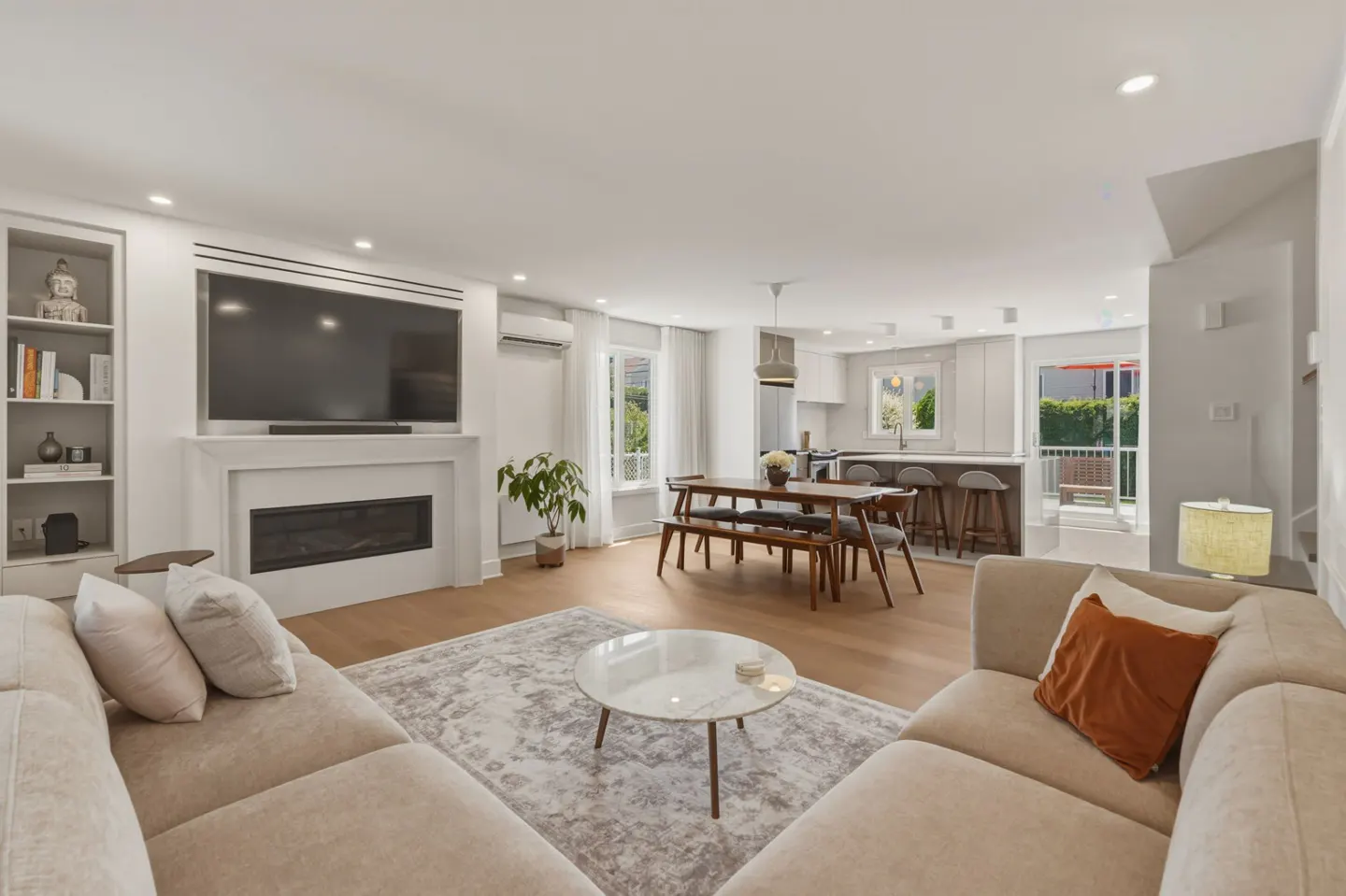 Modern open-plan living room with beige sofas, a fireplace, TV, dining area, and kitchen with a breakfast bar.