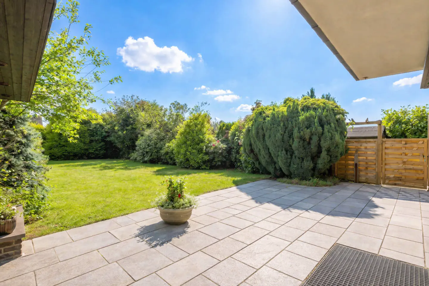 A backyard with a stone patio, green lawn, trees, and a blue sky with white clouds.
