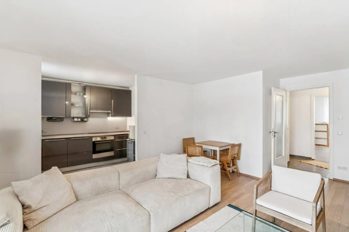 Bright, open-concept apartment with a cream sofa, white walls, and wood floors. A modern kitchen and dining area are visible in the background.