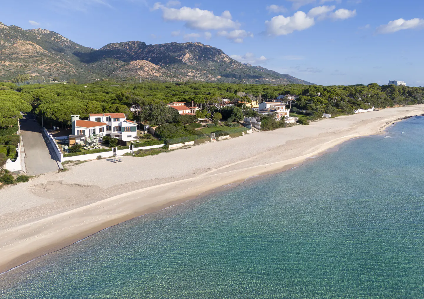 Aerial view of a white beachfront house with a red roof, sandy beach, turquoise water, and green trees. Mountains are in the background.