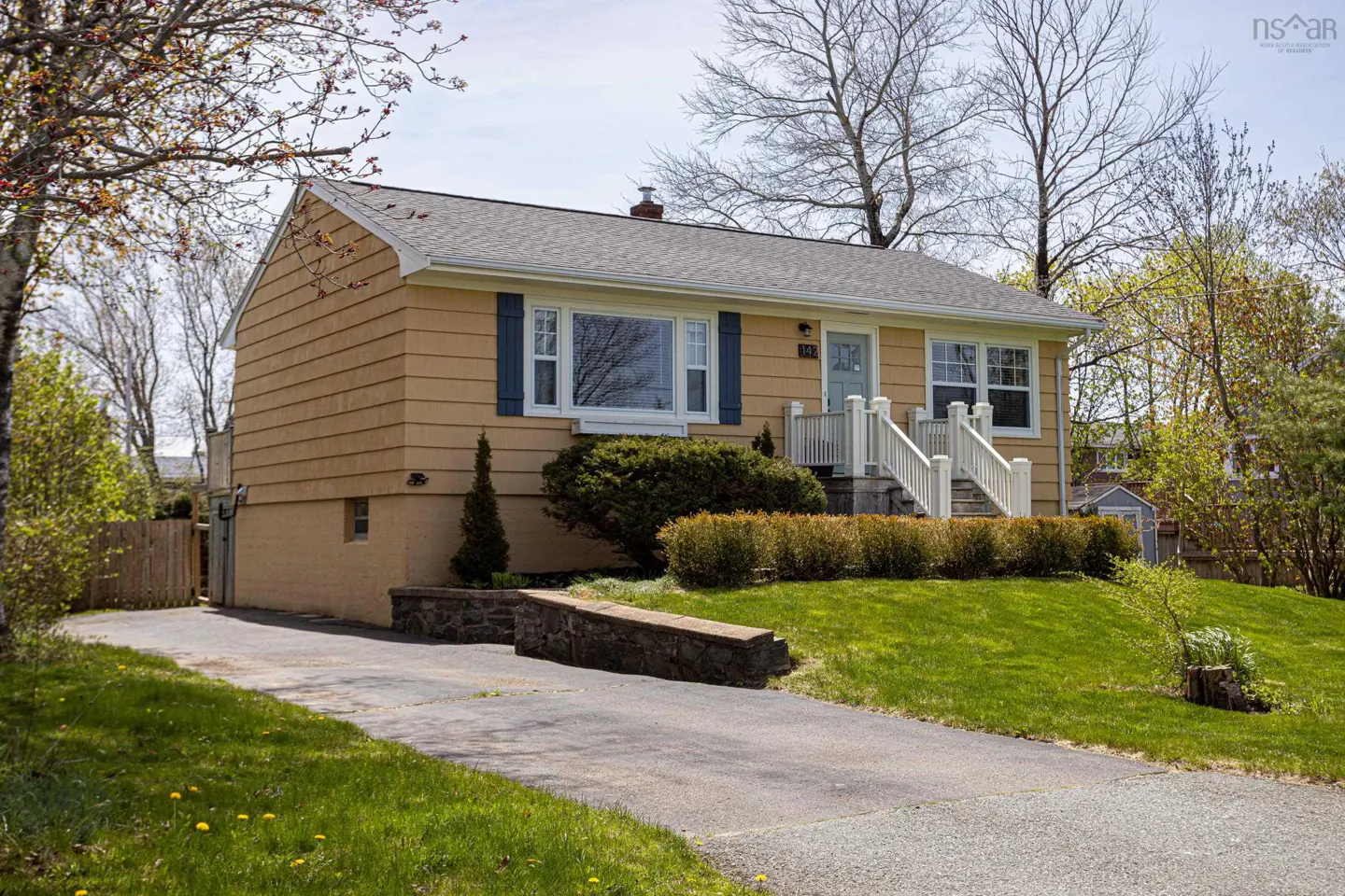 A tan house with blue shutters and a gray roof sits on a green lawn with a paved driveway.