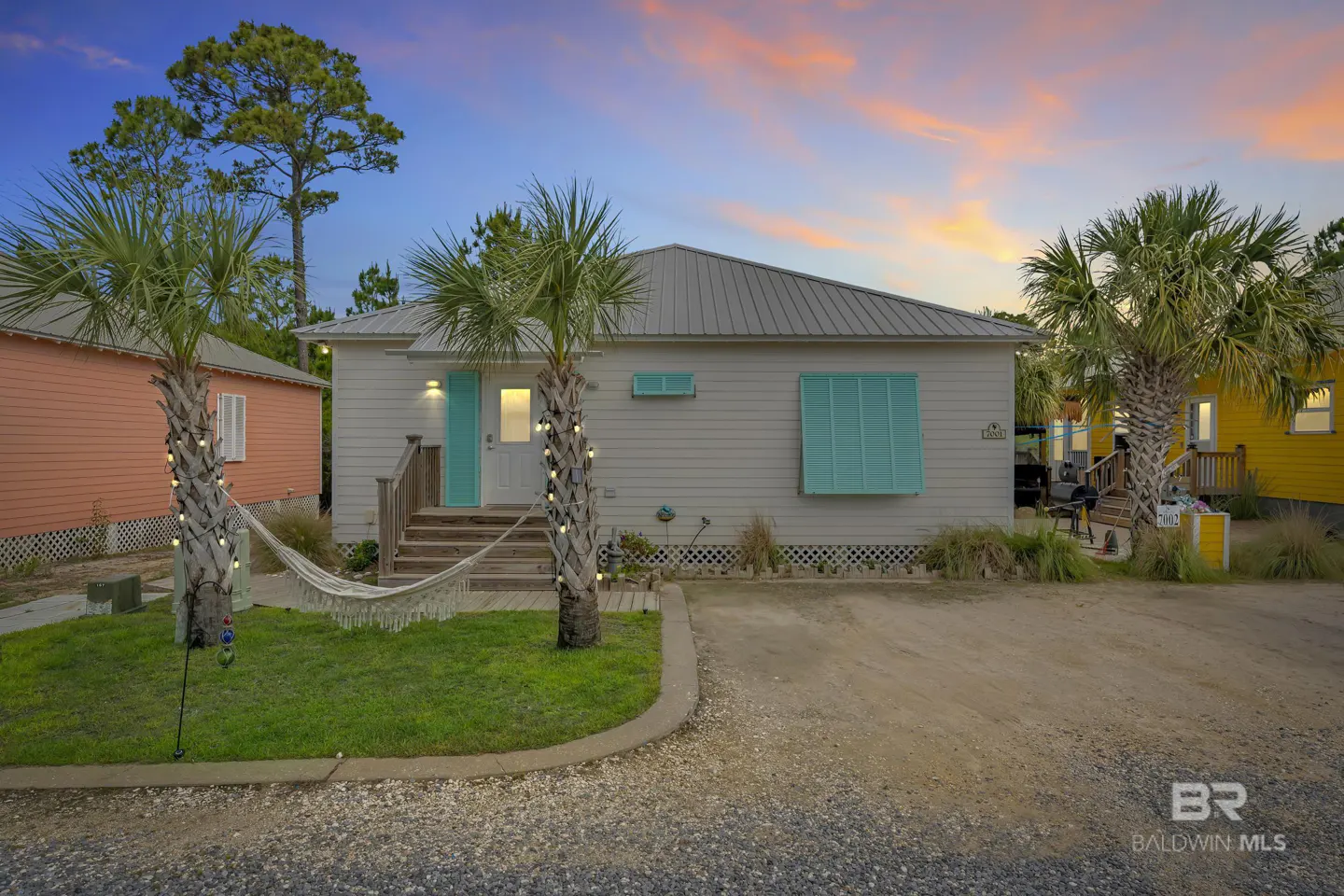 Exterior of a light gray beach house with turquoise shutters and door. Palm trees flank the entrance, and a hammock hangs in the yard.