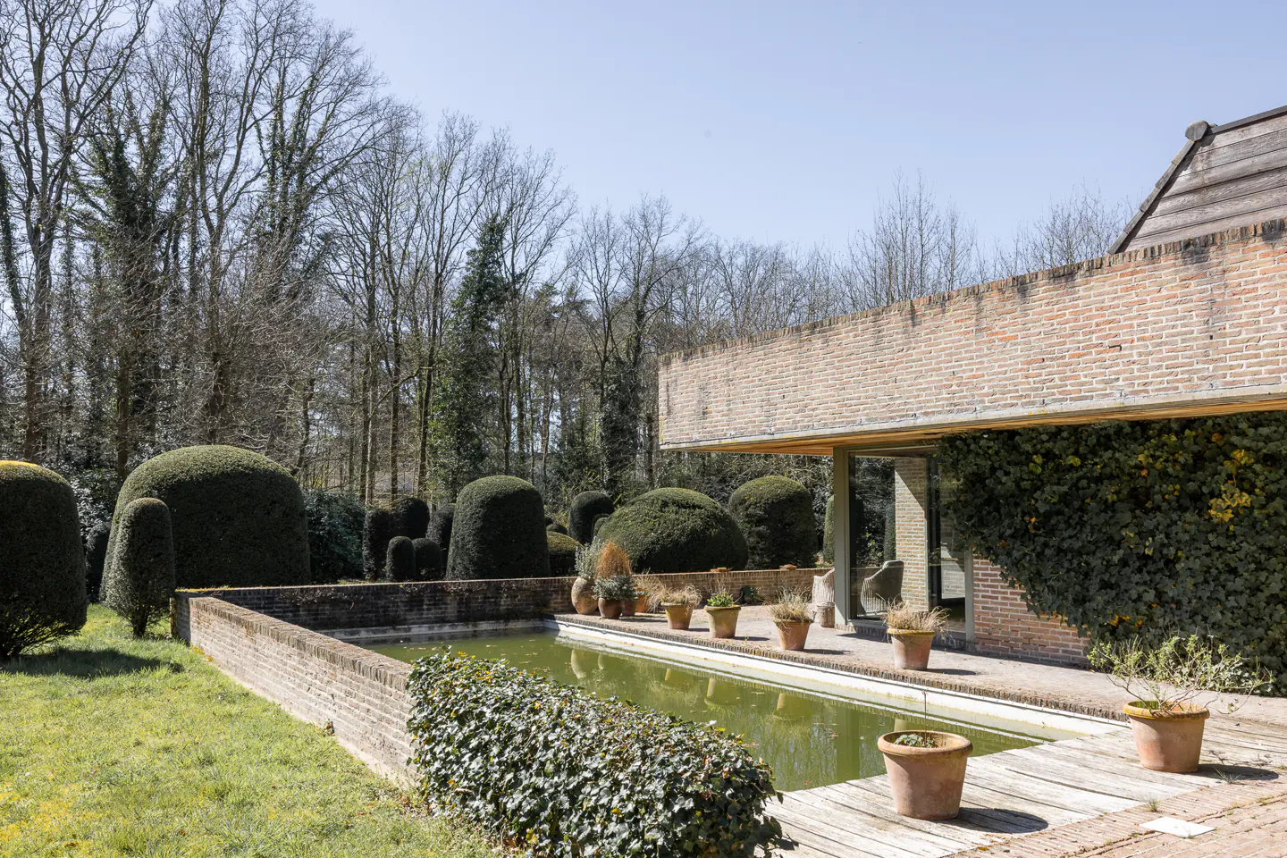 Brick home with a green pool, potted plants, and sculpted bushes. Trees are in the background.
