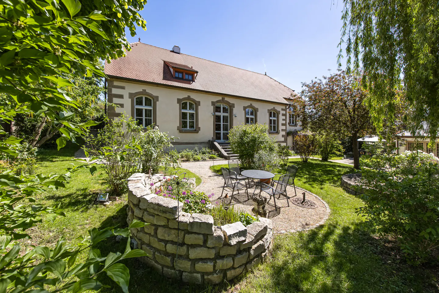 Exterior of a cream-colored house with a red roof, surrounded by a green lawn, trees, and a stone-walled flower bed. A patio set sits on a gravel area.