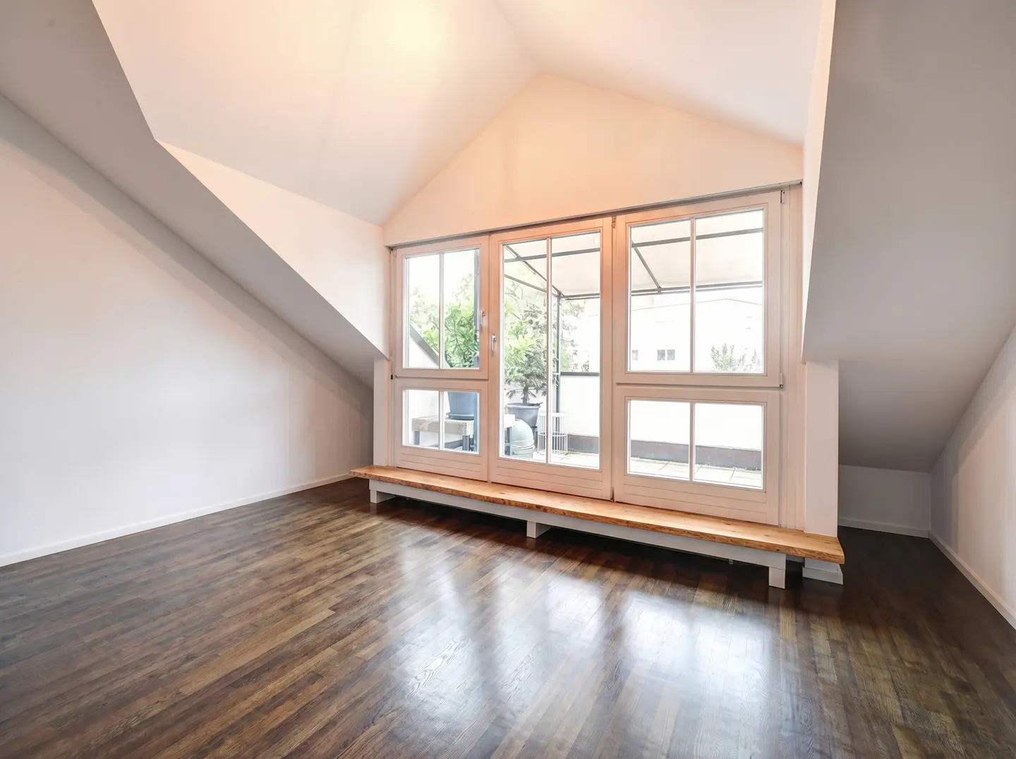 Attic room with dark wood floors, white walls, and a large window with a view of a patio.