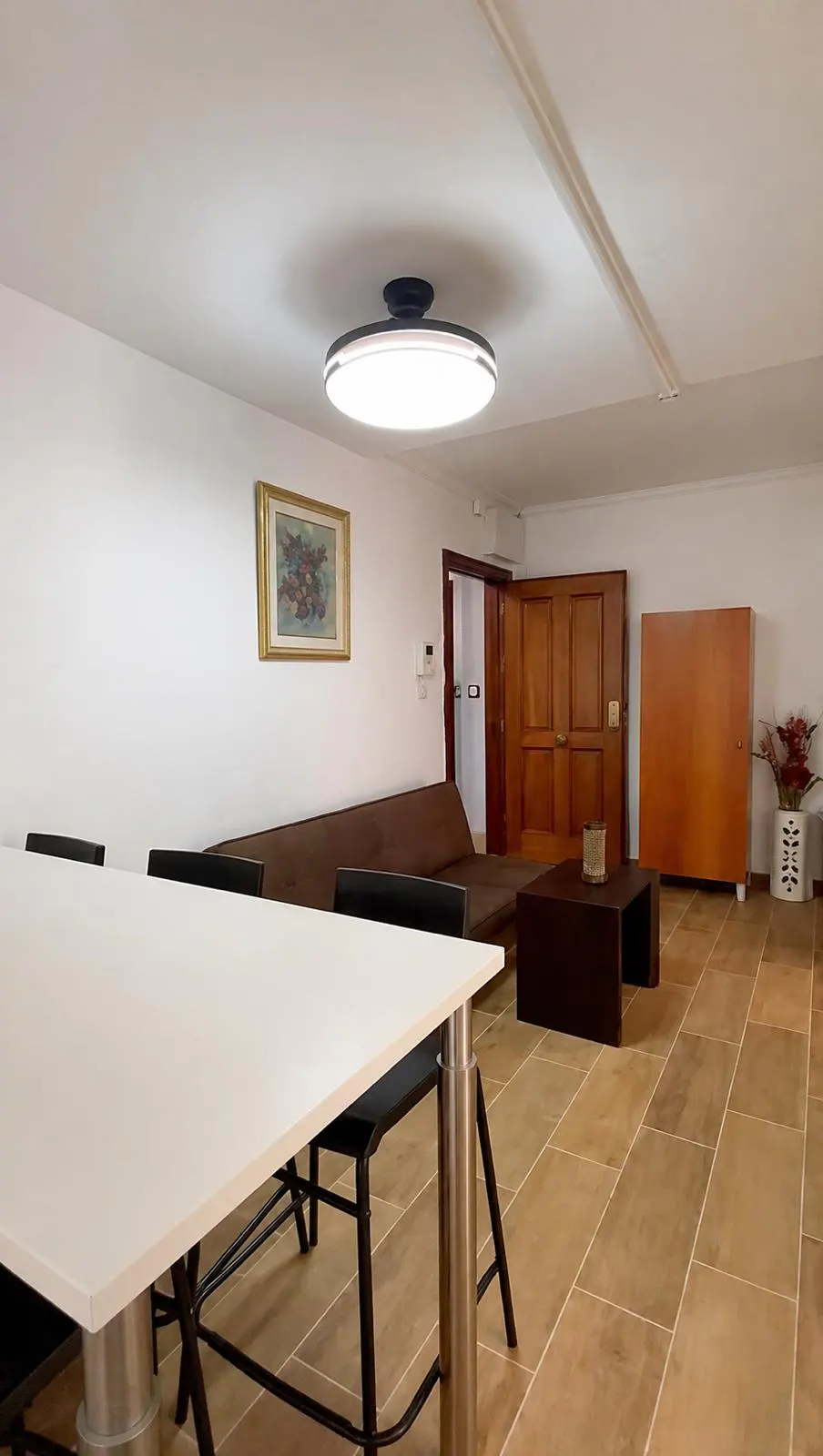 A living room with a white table, black chairs, a brown sofa, and a brown wooden cabinet. The floor is tiled in beige.