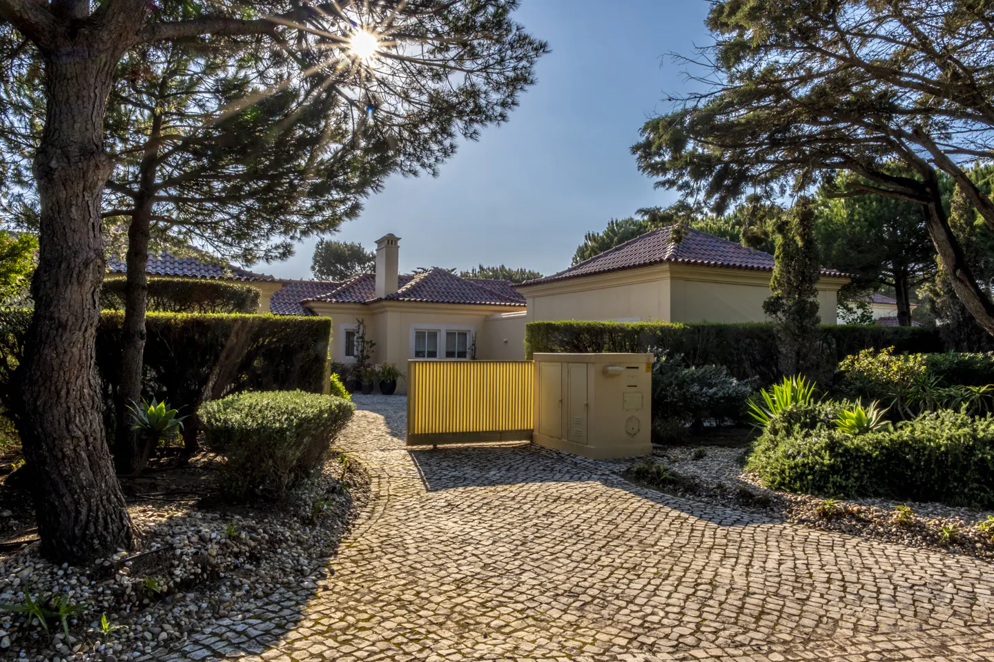 Exterior view of a tan house with a red tile roof, yellow gate, and cobblestone driveway. Trees frame the house.