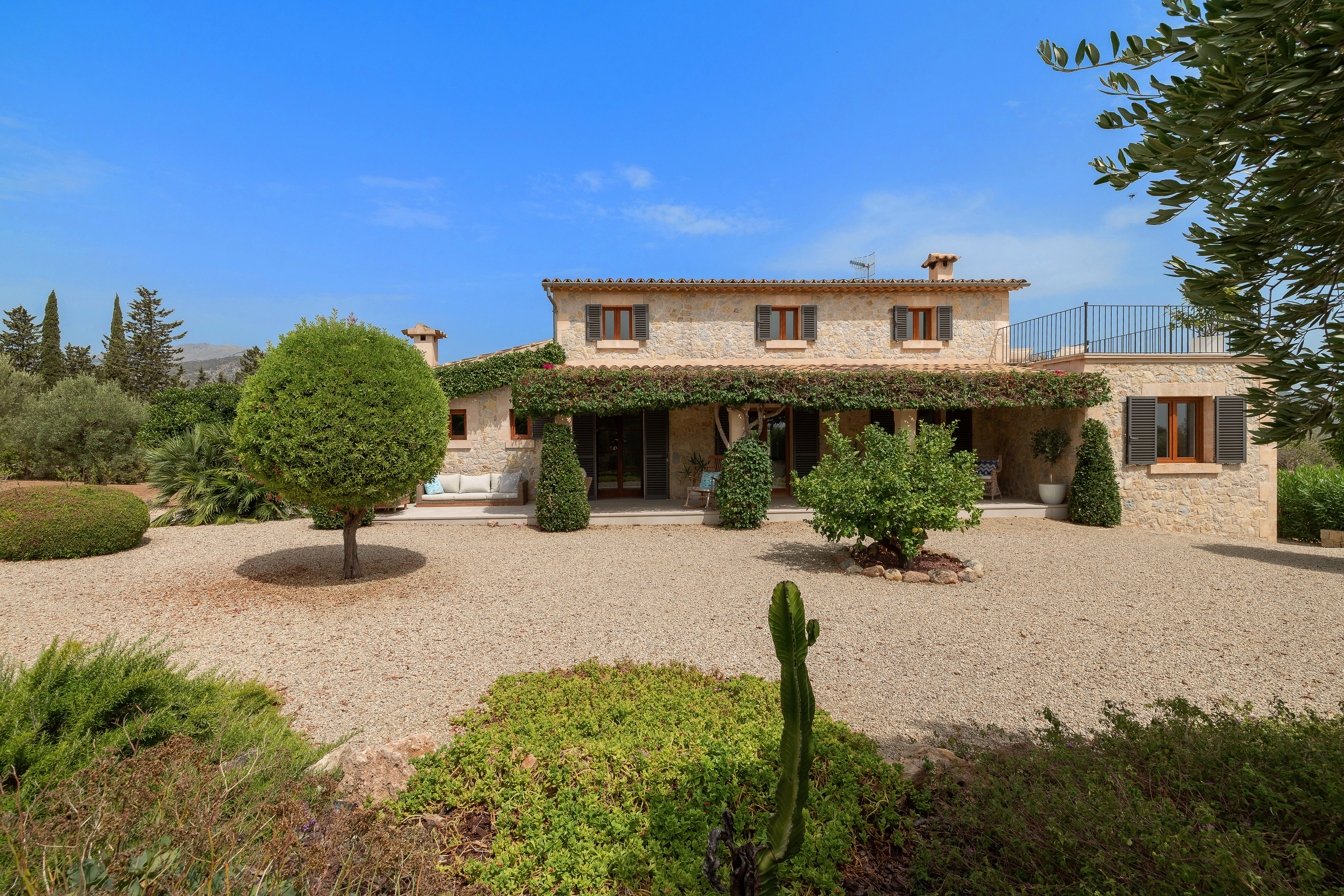 A stone Mediterranean villa with a gravel courtyard, manicured shrubs, and a vine-covered porch under a clear blue sky.