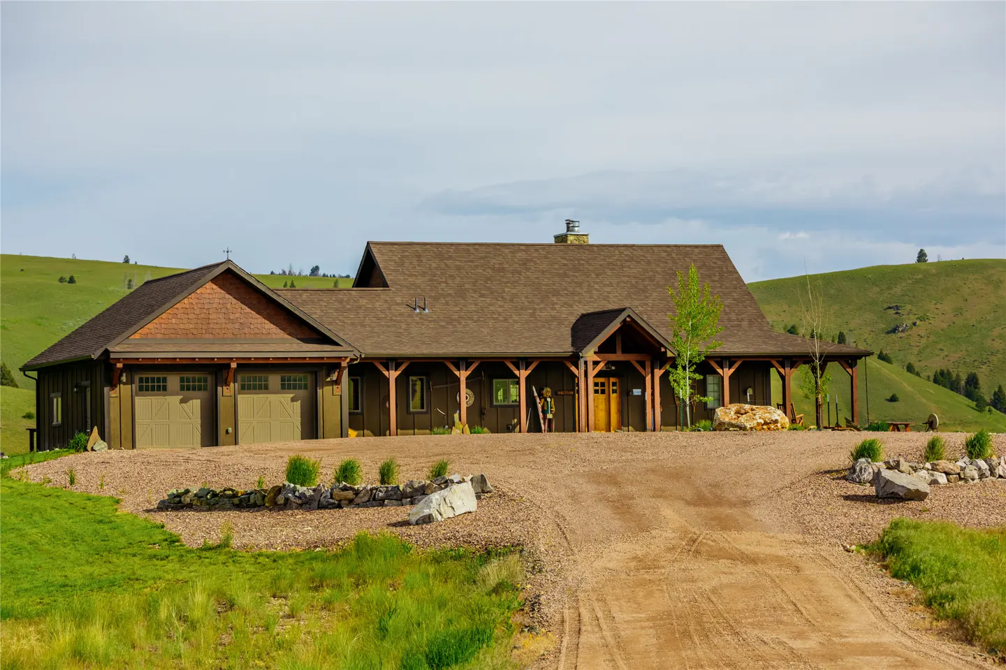 A brown ranch-style home with a two-car garage sits at the end of a dirt driveway, surrounded by rolling green hills.