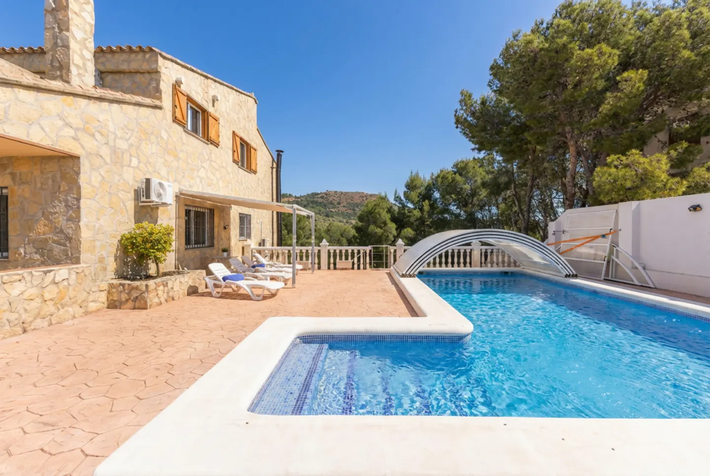 Exterior view of a stone house with a pool, lounge chairs, and a retractable pool cover on a sunny day.