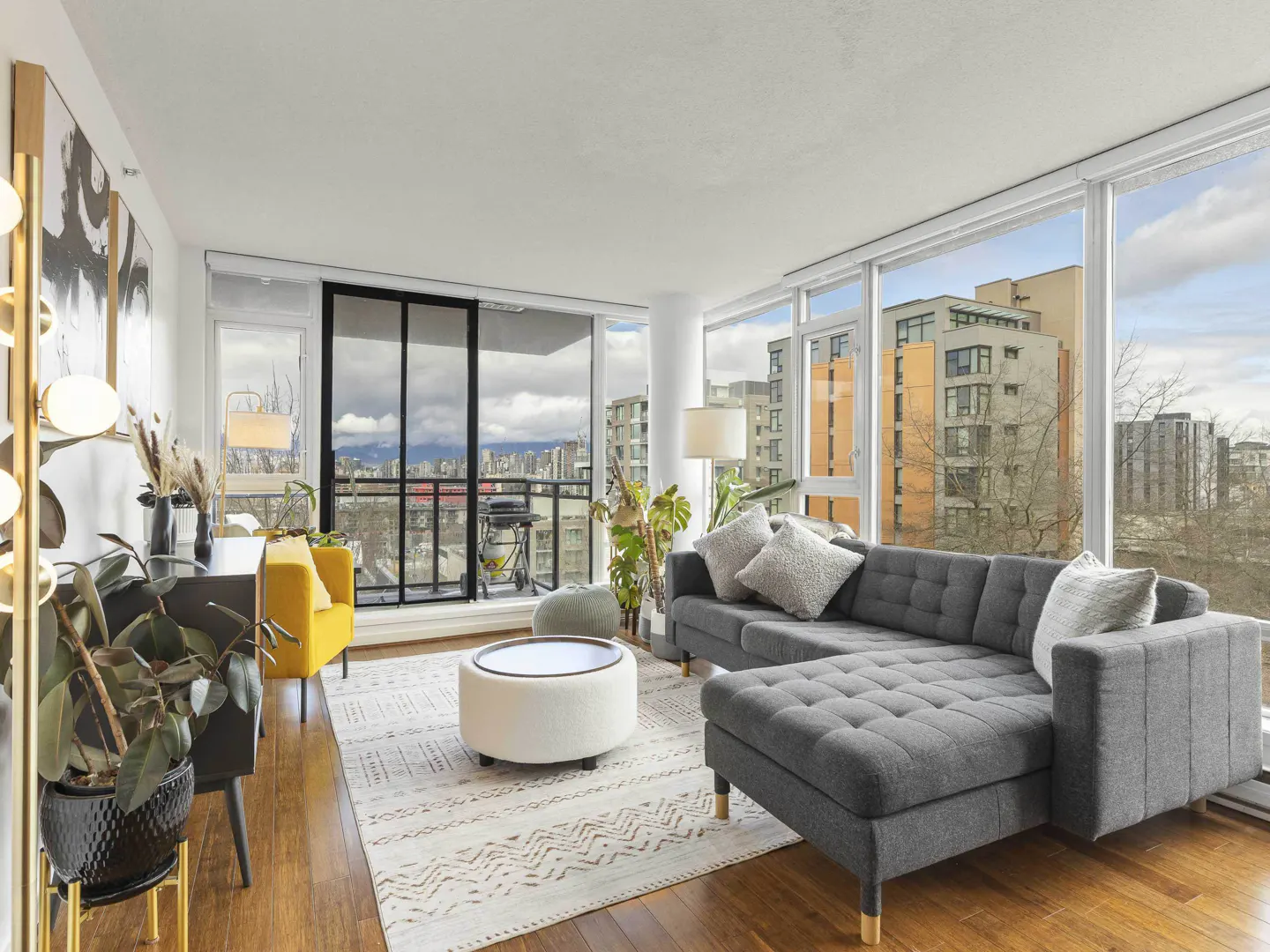 Bright living room with a gray sectional, yellow chair, and city view. Hardwood floors and a patterned rug add warmth.