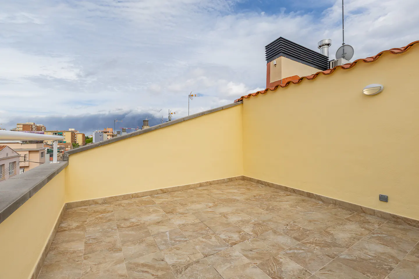 Empty rooftop terrace with beige tiled floor, yellow walls, and a view of the city under a cloudy sky.