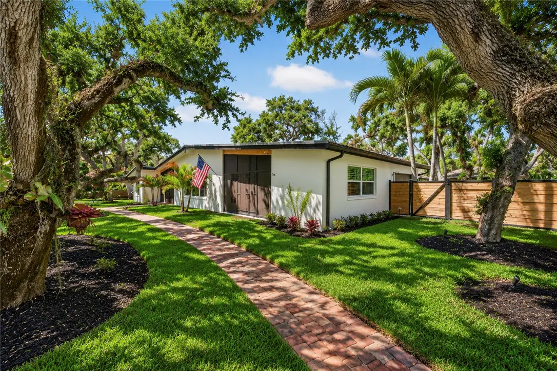 A white house with a brick path leading to the front door, an American flag, and lush green trees.