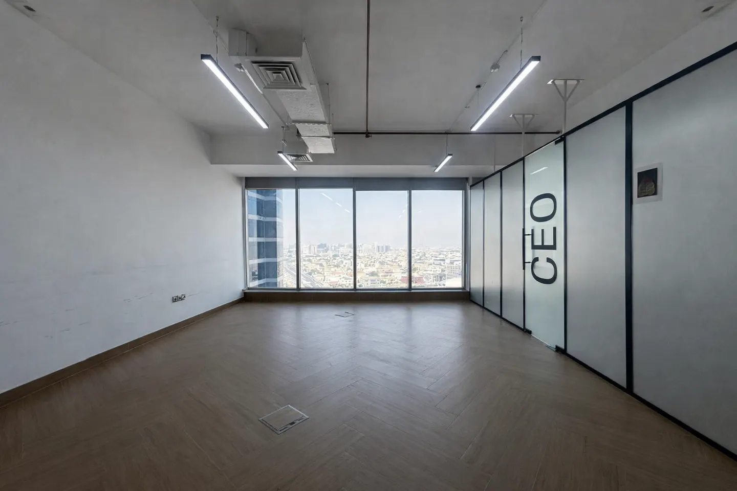Empty office space with wood floors, white walls, and a large window overlooking a city. "CEO" is printed on a glass wall.