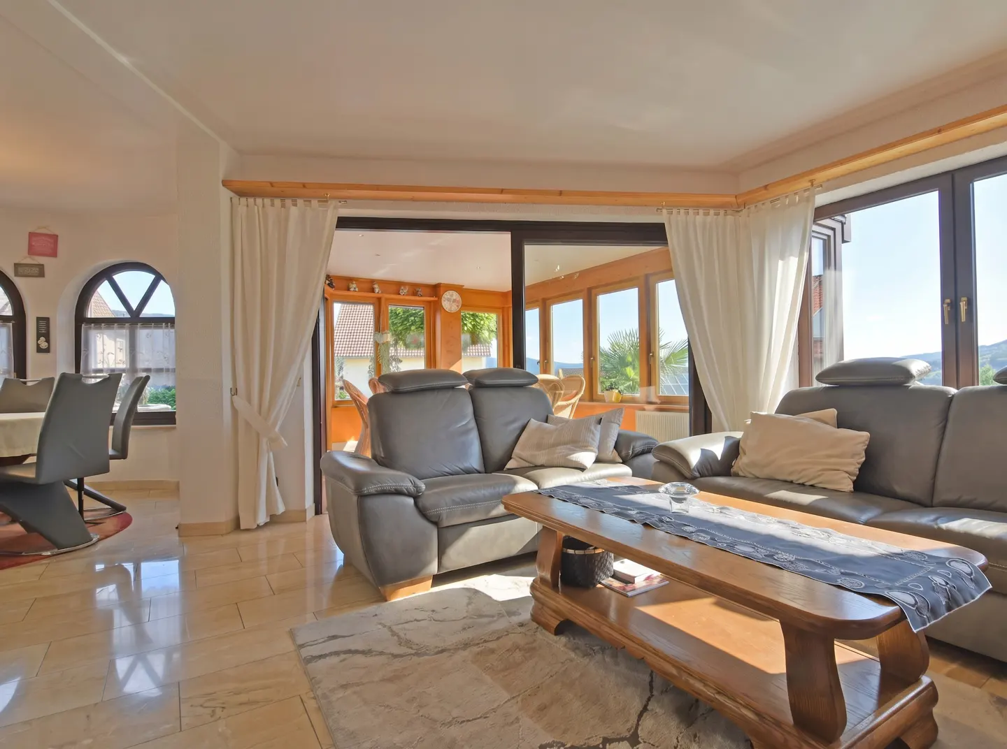 Living room with gray leather sofas, wood coffee table, and marble floors. Dining area visible through arched doorway. Sunroom in background.