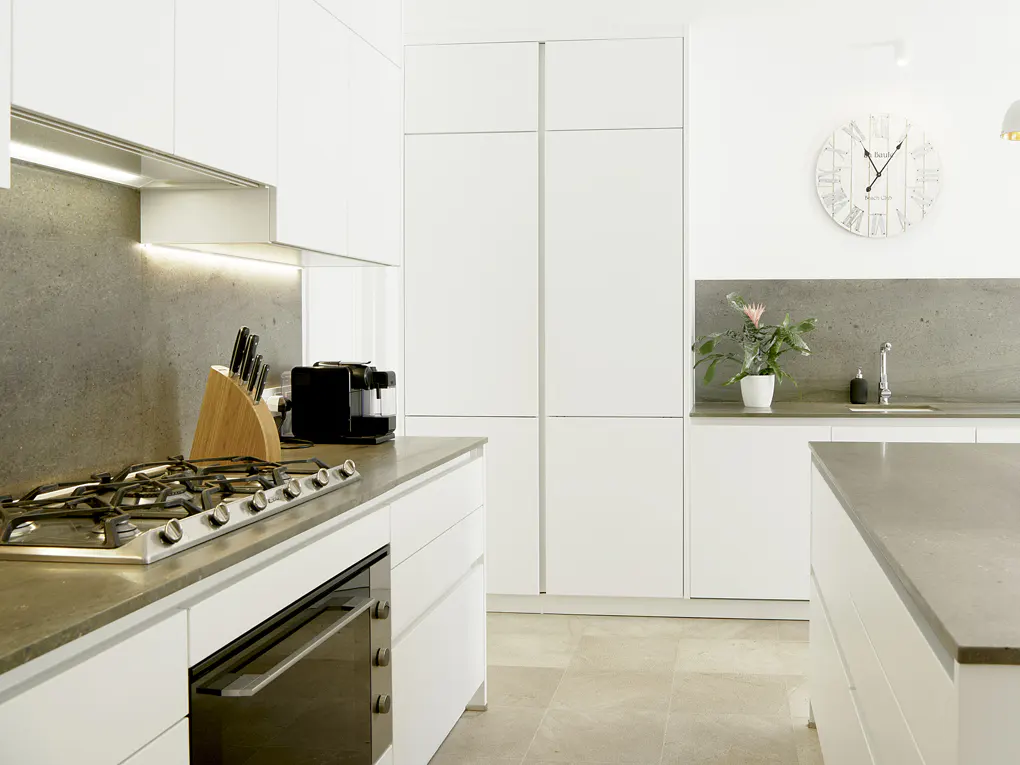 Bright kitchen with white cabinets, gray countertops, and stainless steel appliances. A clock hangs on the wall above a sink and a potted plant.