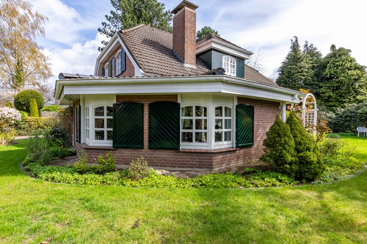 Exterior view of a brick house with green shutters, white trim, and a brown tiled roof, surrounded by a green lawn and trees.