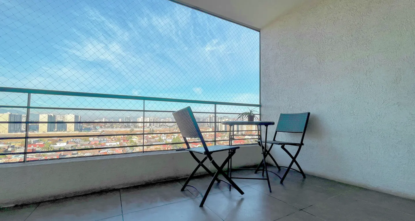 A balcony with two black folding chairs and a small table, overlooking a city skyline under a bright blue sky.