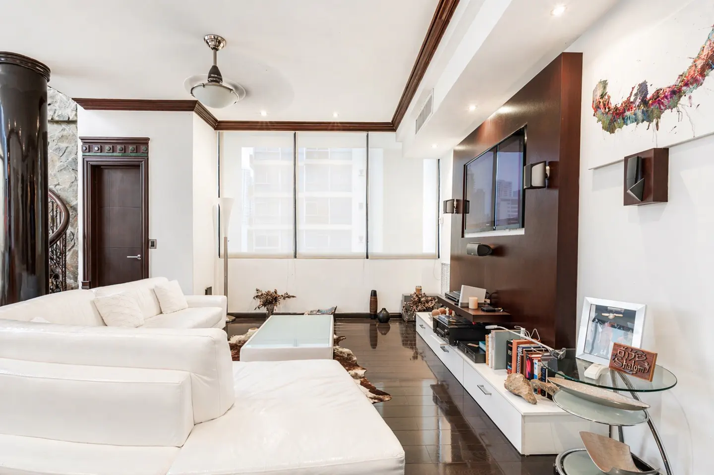 Bright living room with white sofas, dark wood floors, and a large TV mounted on a dark wood panel. A glass coffee table sits in front of the sofas.