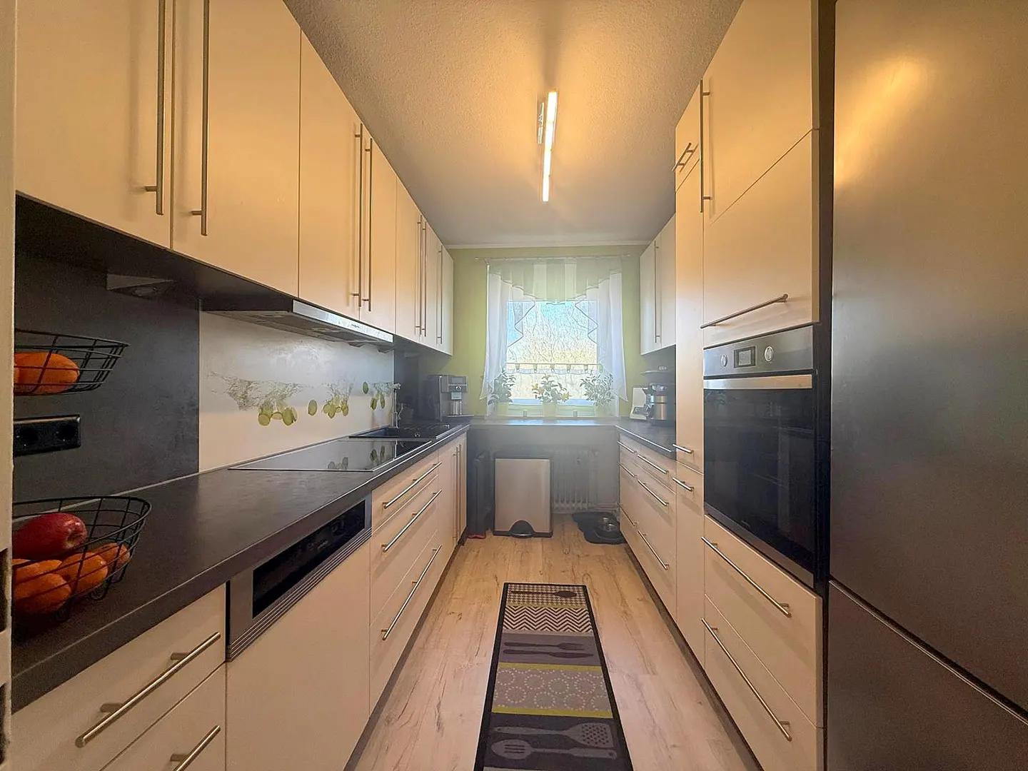 A galley kitchen with light wood cabinets, dark countertops, and stainless steel appliances. A window with white curtains is at the end of the kitchen.