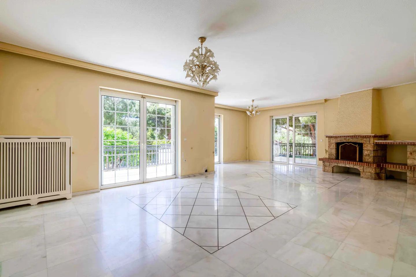 Bright living room with marble floors, yellow walls, and white trim. French doors lead to balconies. A brick fireplace and chandelier add detail.