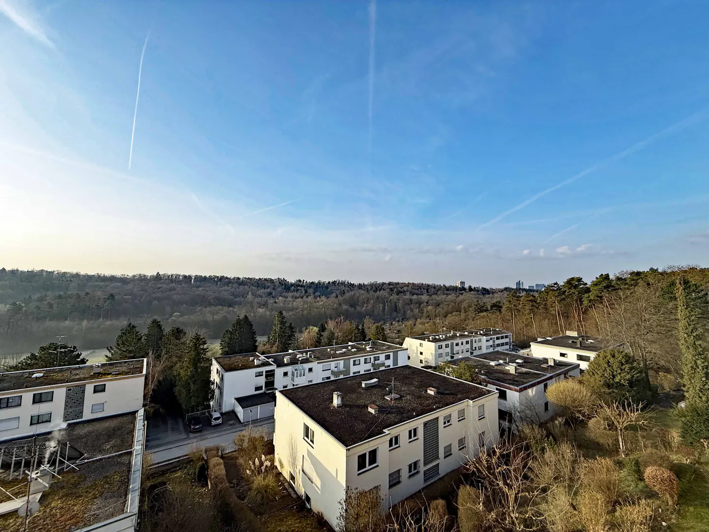 View of white apartment buildings with dark roofs, surrounded by trees under a blue sky with contrails.