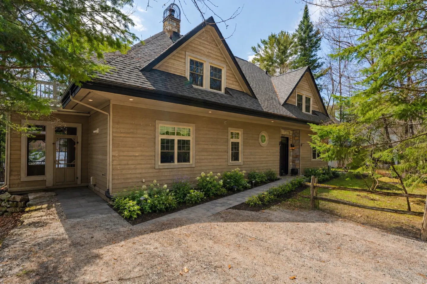 Exterior view of a tan house with a black roof, surrounded by trees and greenery. A stone walkway leads to the front door.