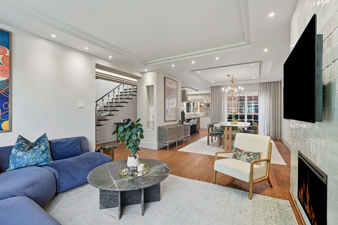 Open-concept living room with blue sofa, marble table, fireplace, and dining area. Staircase visible in the background.