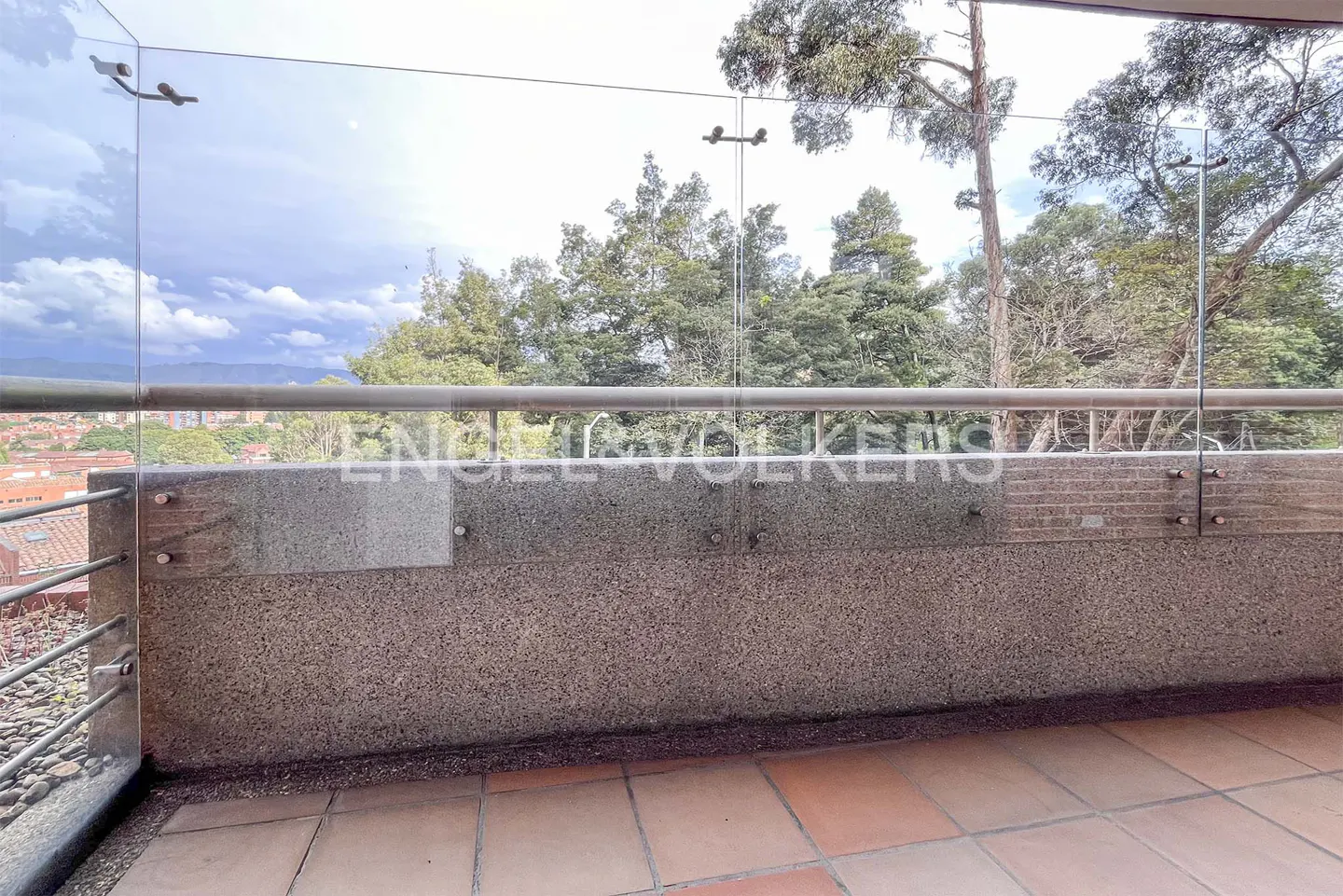Balcony view with glass railing, stone wall, and terracotta tiles. Trees and city buildings are visible in the background.