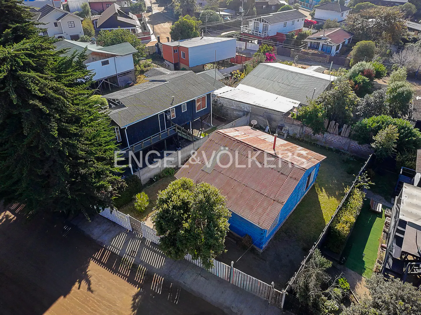 Aerial view of a blue house with a rusty red roof, surrounded by a white picket fence and green trees in a residential area.