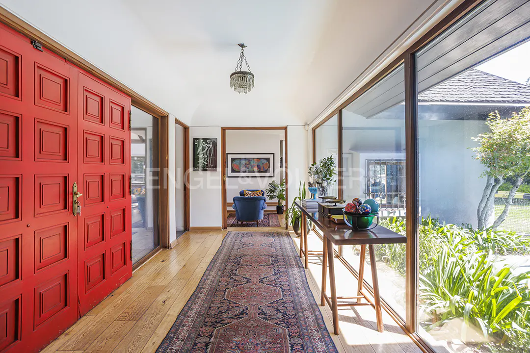 Bright entryway with a red door, wood floors, and a long rug. A table sits by large windows overlooking greenery. Chandelier hangs from the white ceiling.