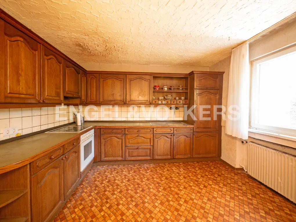 A dated kitchen with wood cabinets, brown tile floor, white oven, and a window with a white curtain.