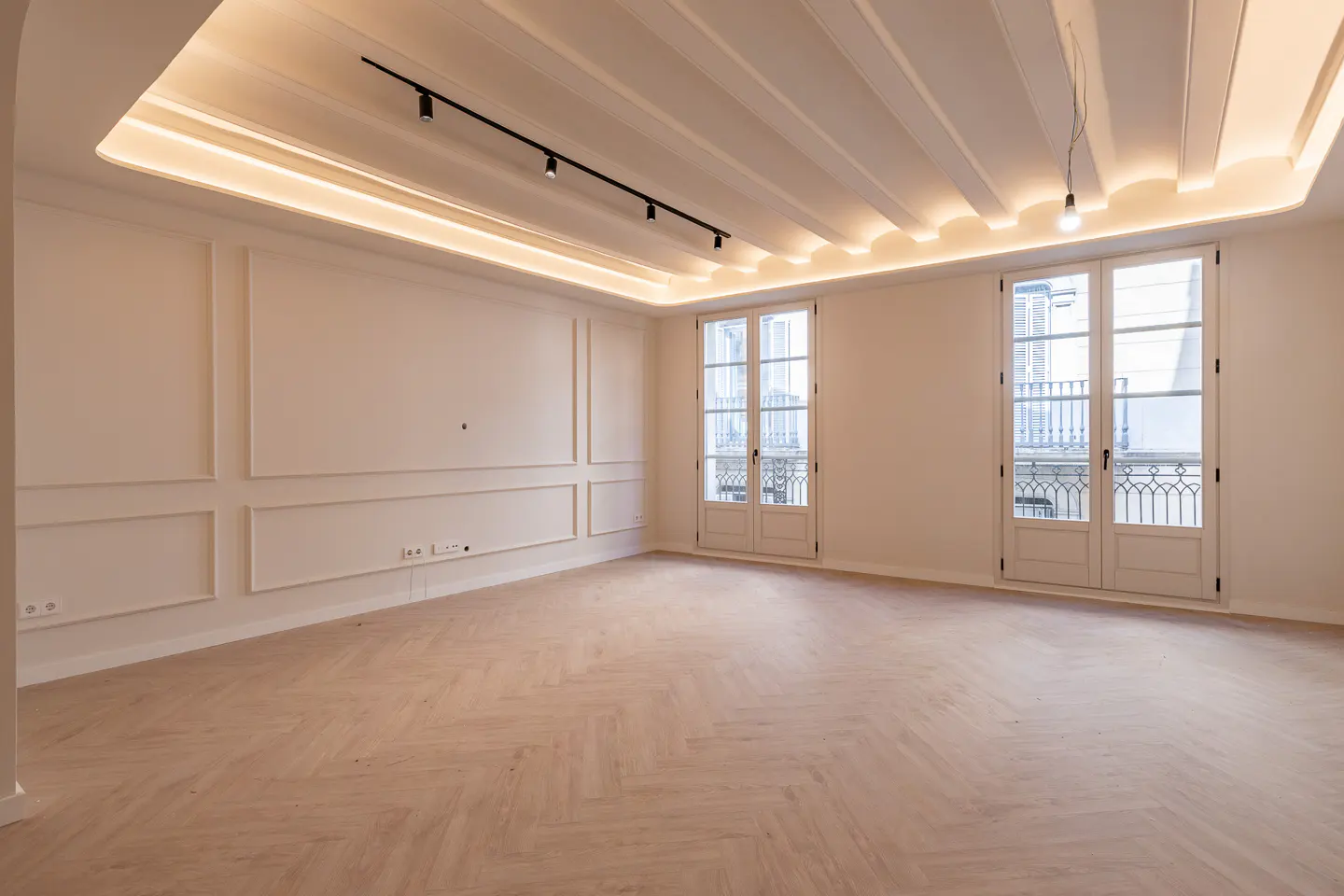 Bright, empty room with herringbone wood floors, white walls with molding, and two sets of French doors. Track lighting and recessed lighting illuminate the space.