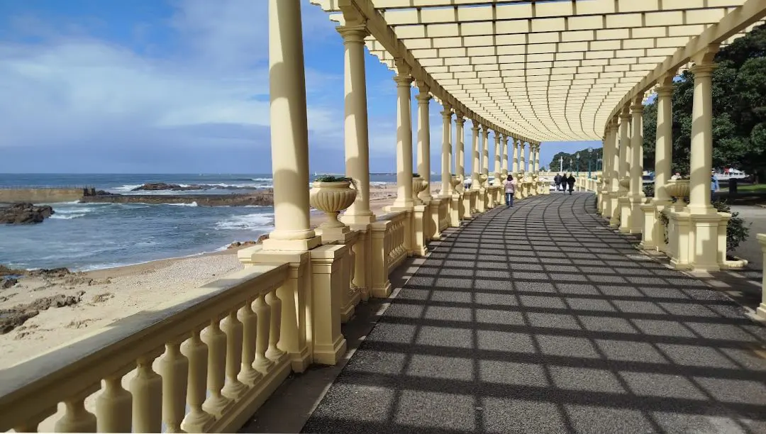 Coastal walkway with a cream-colored pergola, columns, and balustrade. Ocean view with rocks and beach to the left. People walking in the distance.