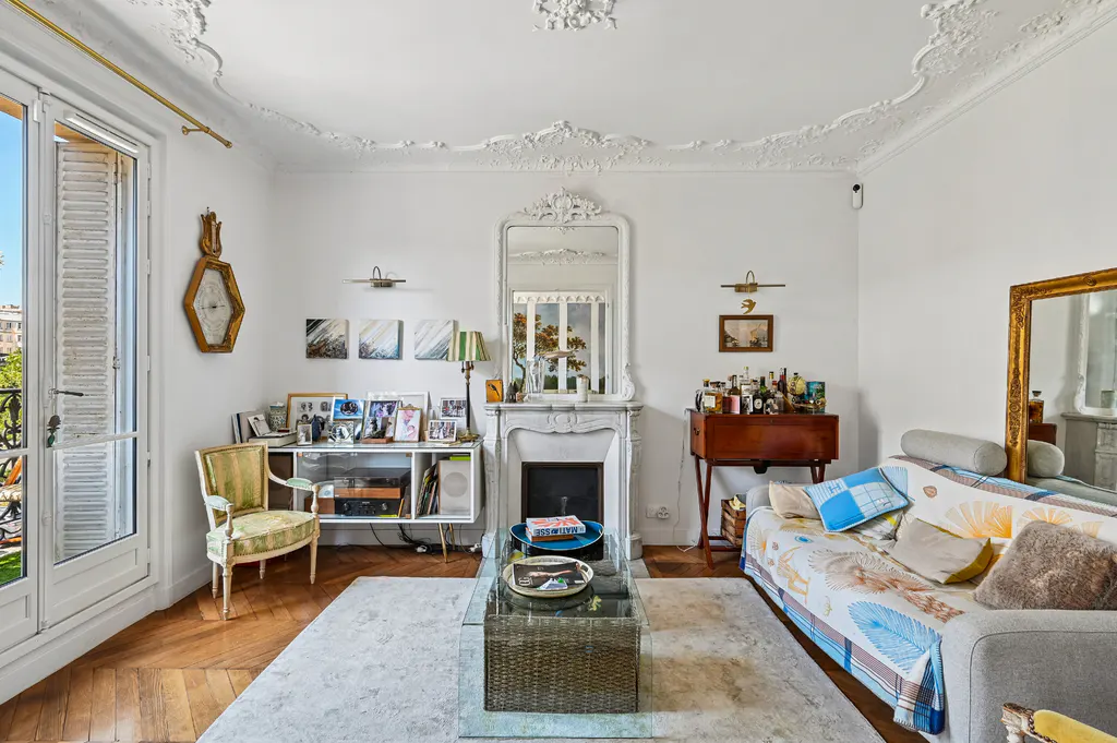 Bright living room with white walls, herringbone wood floors, and ornate ceiling molding. A fireplace, sofa, and glass coffee table furnish the space.