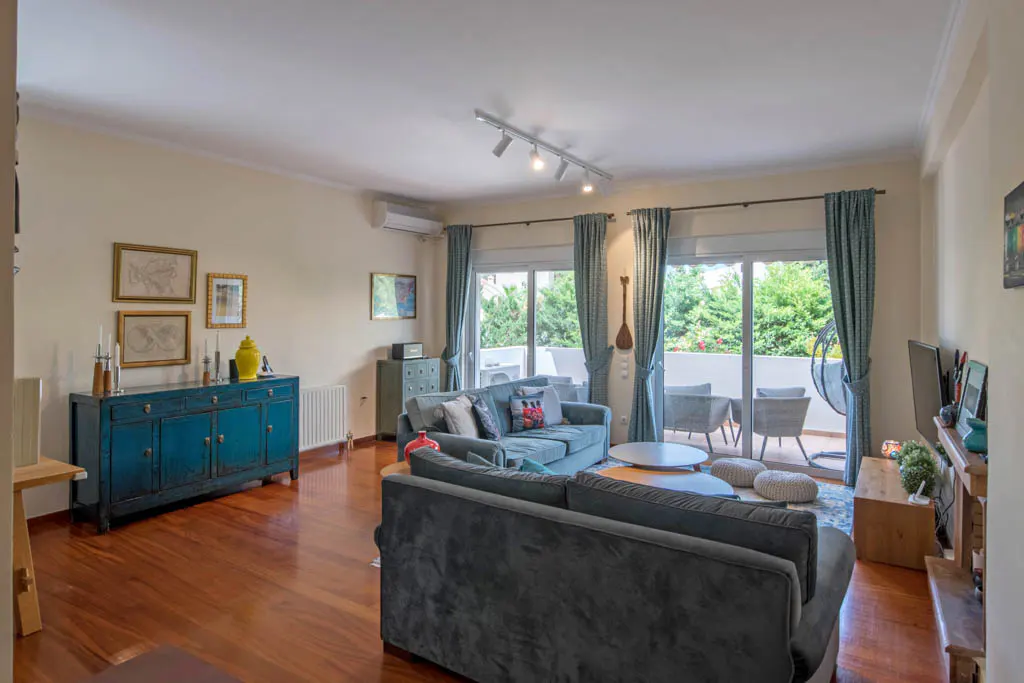 A living room with hardwood floors, blue sofas, and a blue cabinet with framed art above. Sliding glass doors lead to a balcony.