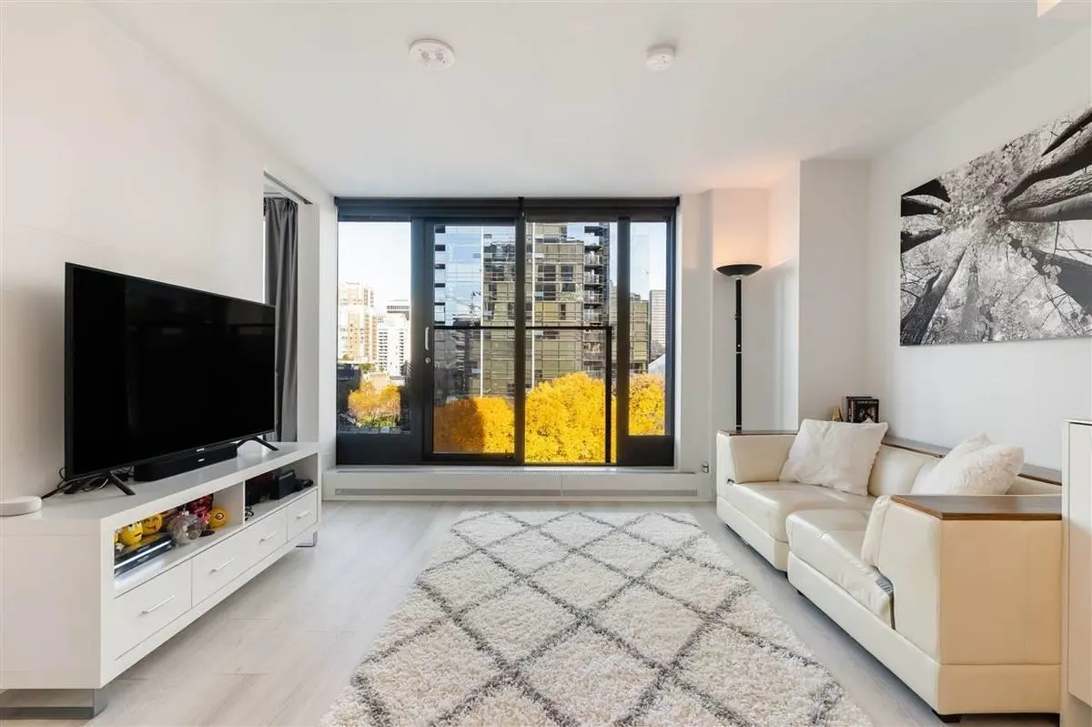 Bright living room with white walls, a large window showing city views, a white sofa, and a TV on a white stand.
