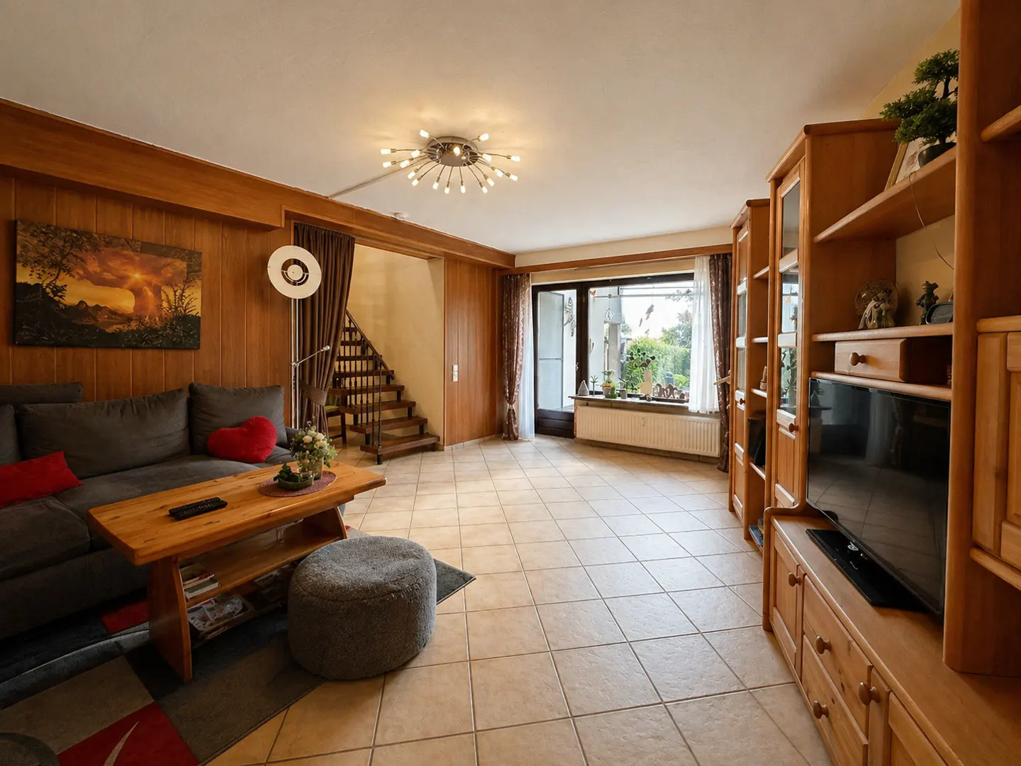Living room with wood paneling, tile floor, gray sofa, wood table, and built-in wood shelving with a TV. A staircase is visible.