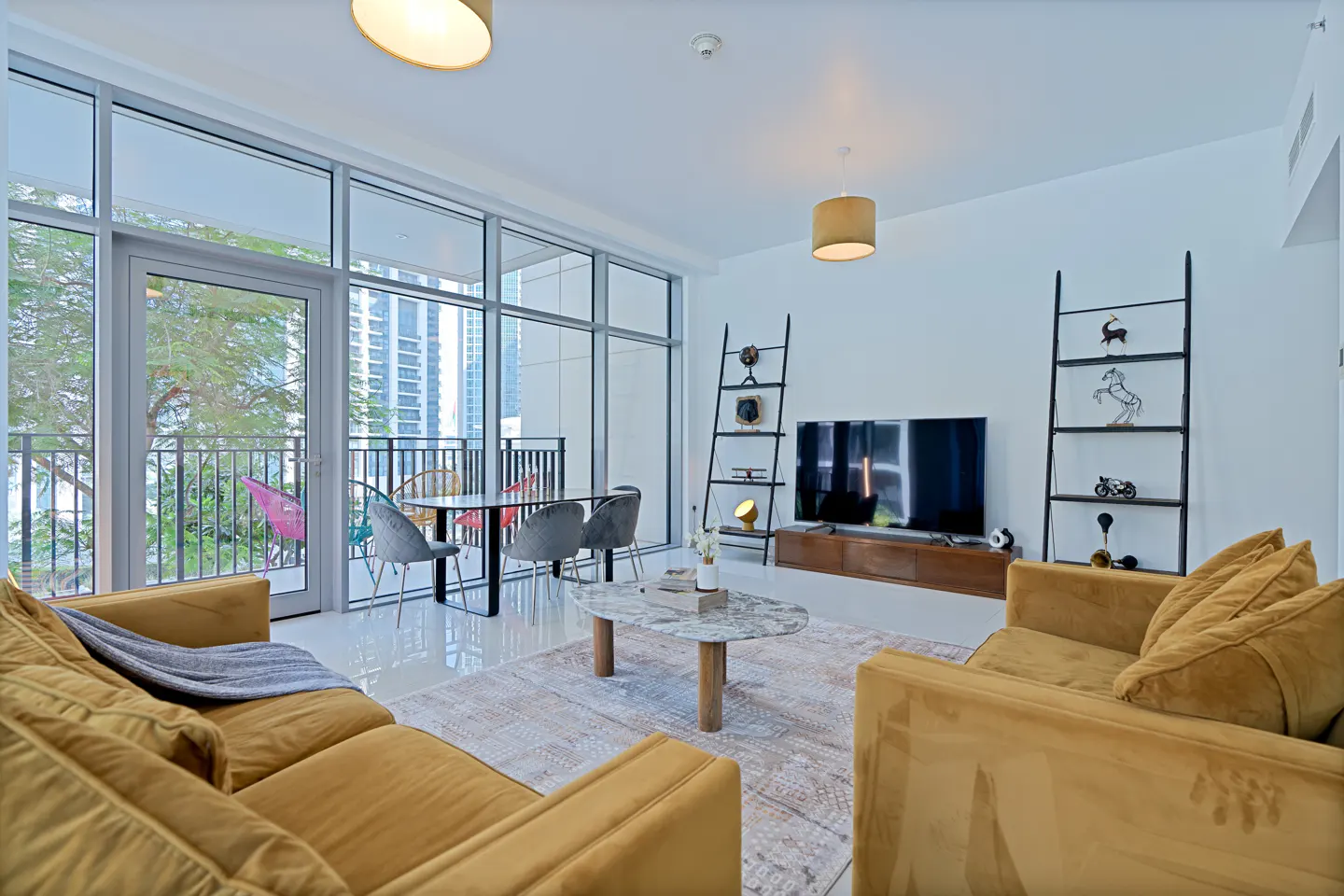 Bright living room with yellow sofas, a marble coffee table, and a balcony with city views. Black ladder shelves flank a TV.