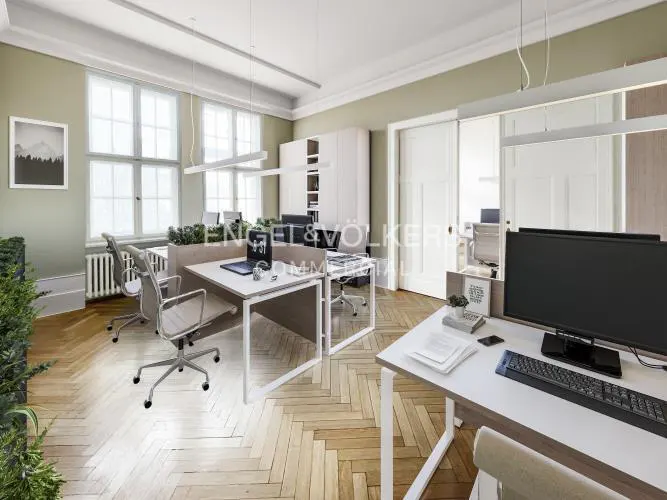 Bright office space with desks, chairs, computers, and herringbone wood floors. Large windows and white cabinets are visible.