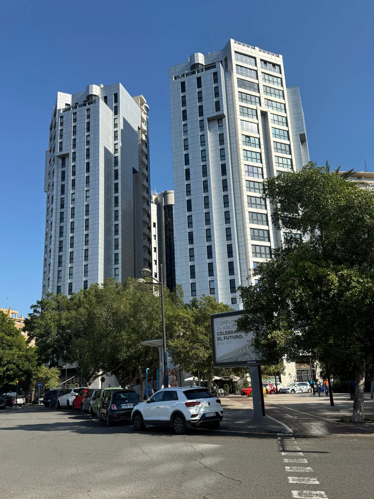 Two tall, modern, silver apartment buildings rise above trees and parked cars on a sunny day with a clear blue sky.