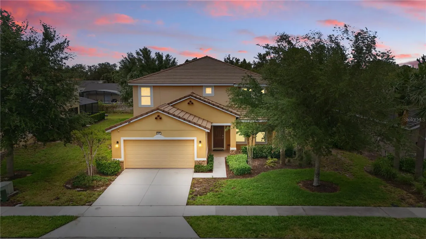Two-story tan house with a brown roof, green lawn, and trees against a pink and blue sky.