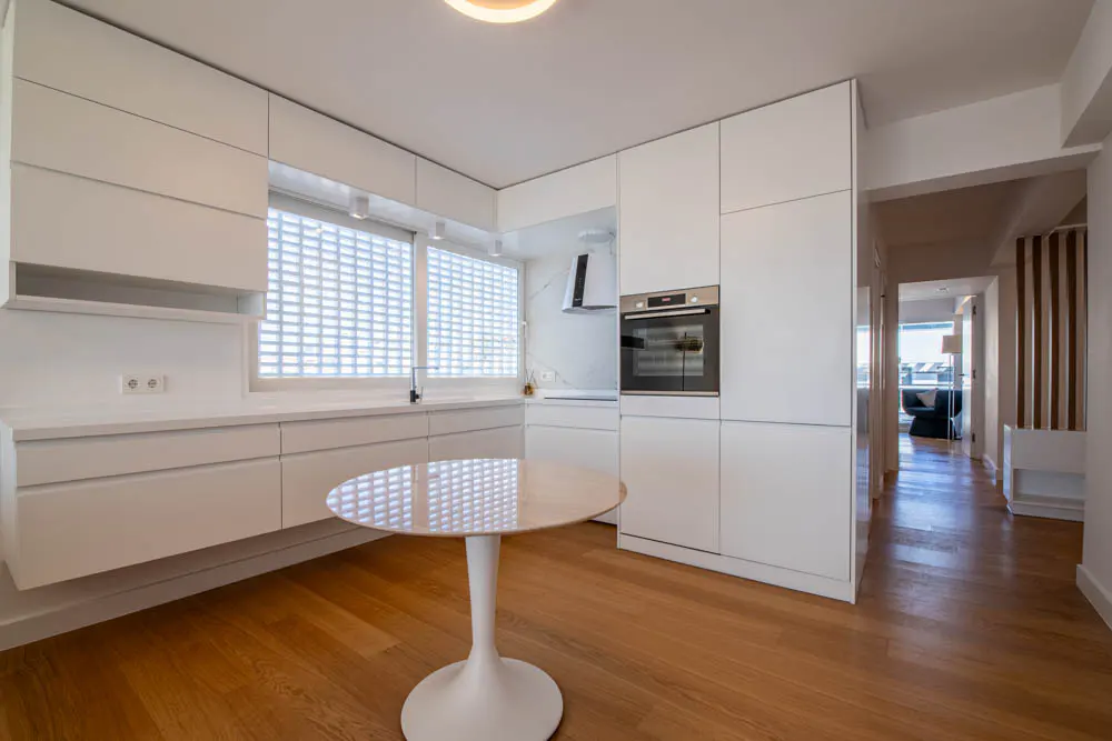 Bright, modern kitchen with white cabinets, wood floors, and a round table. A window with glass blocks provides natural light.