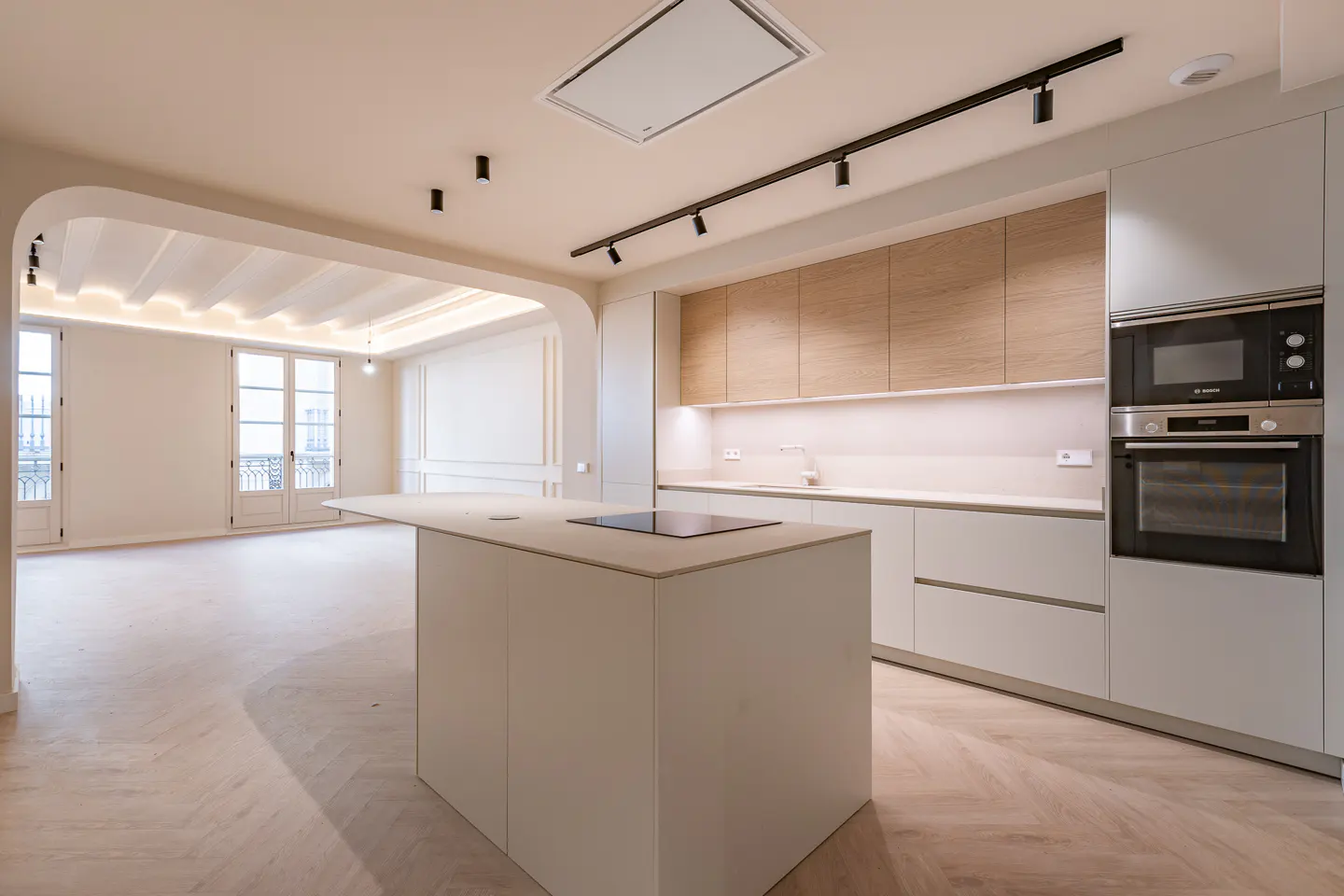 A bright, modern kitchen with white cabinets, wood accents, and an island with a cooktop. The room opens to a living area with French doors.
