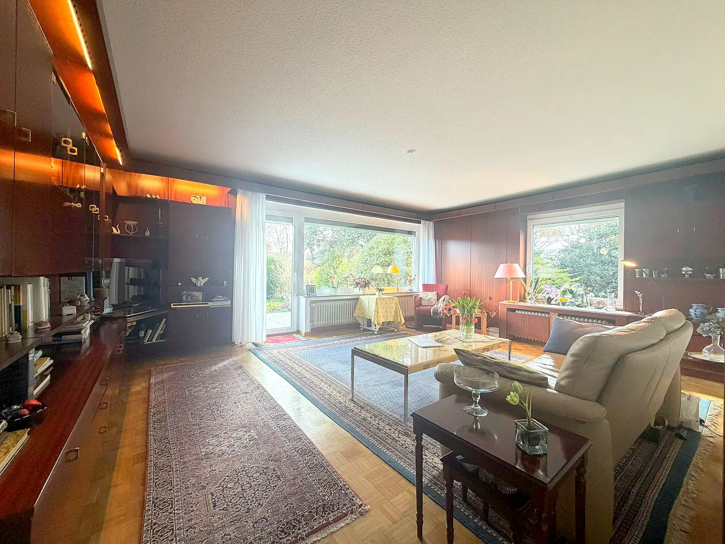 A living room with wood paneled walls, a large window, and a beige sofa. A blue rug covers the wooden floor.