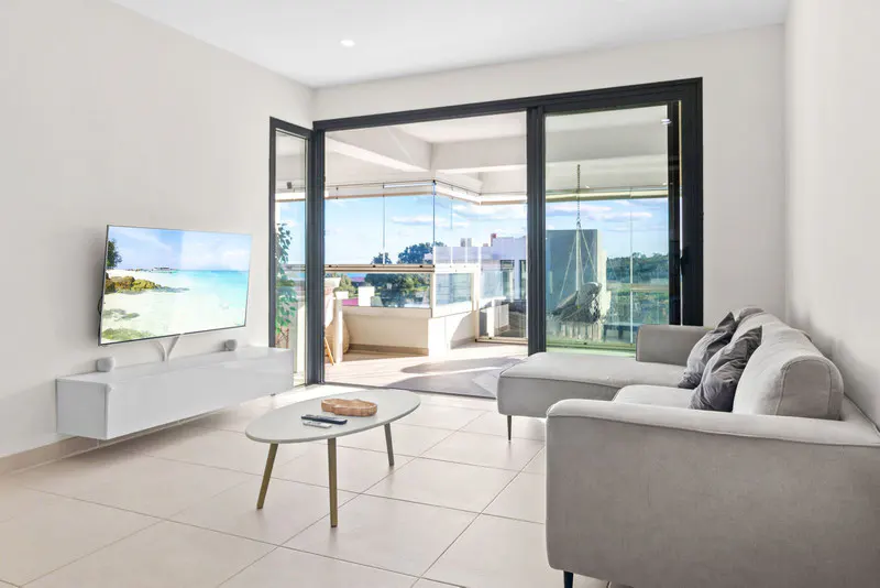 Bright living room with a gray sofa, white walls, and a TV showing a beach scene. Sliding glass doors open to a balcony with a view.