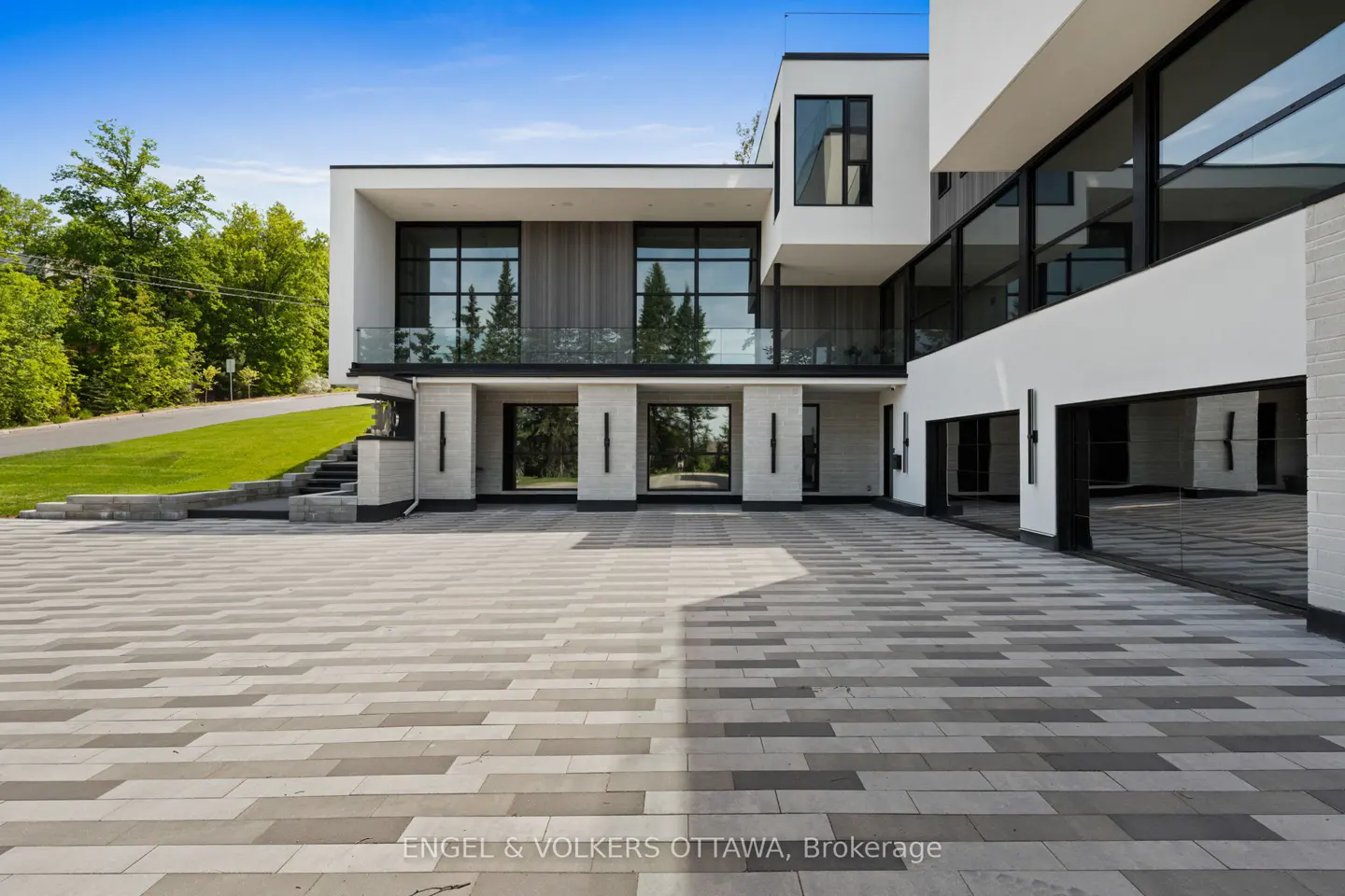 Modern white luxury home with a large paved driveway, glass balcony, and lush green trees under a clear blue sky.