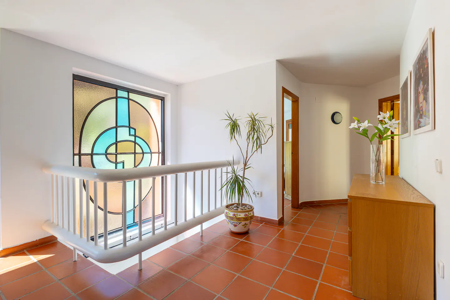 Hallway with terracotta tile floor, white walls, and a stained glass window. A white railing overlooks the lower level. A potted plant and wooden dresser add warmth.
