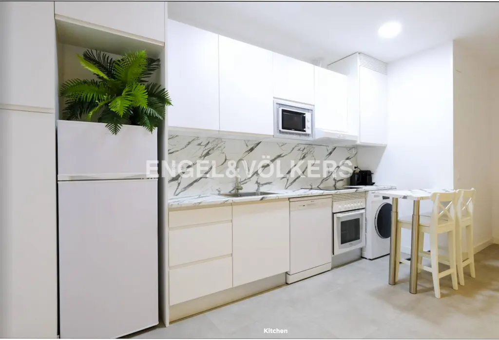 Bright kitchen with white cabinets, marble backsplash, stainless steel appliances, and a small table with two chairs. A green plant sits atop the refrigerator.